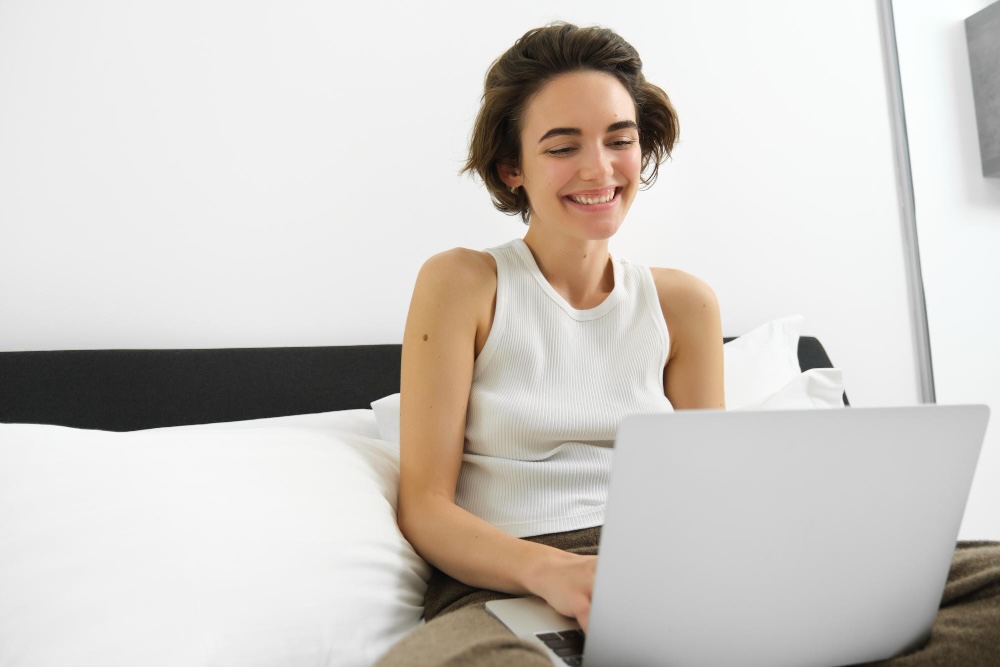 chica joven sentada en la cama sonriendo con el ordenador portátil