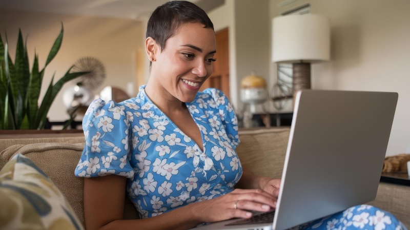 mujer sonriendo en el sofá de su casa tecleando con su ordenador portátil