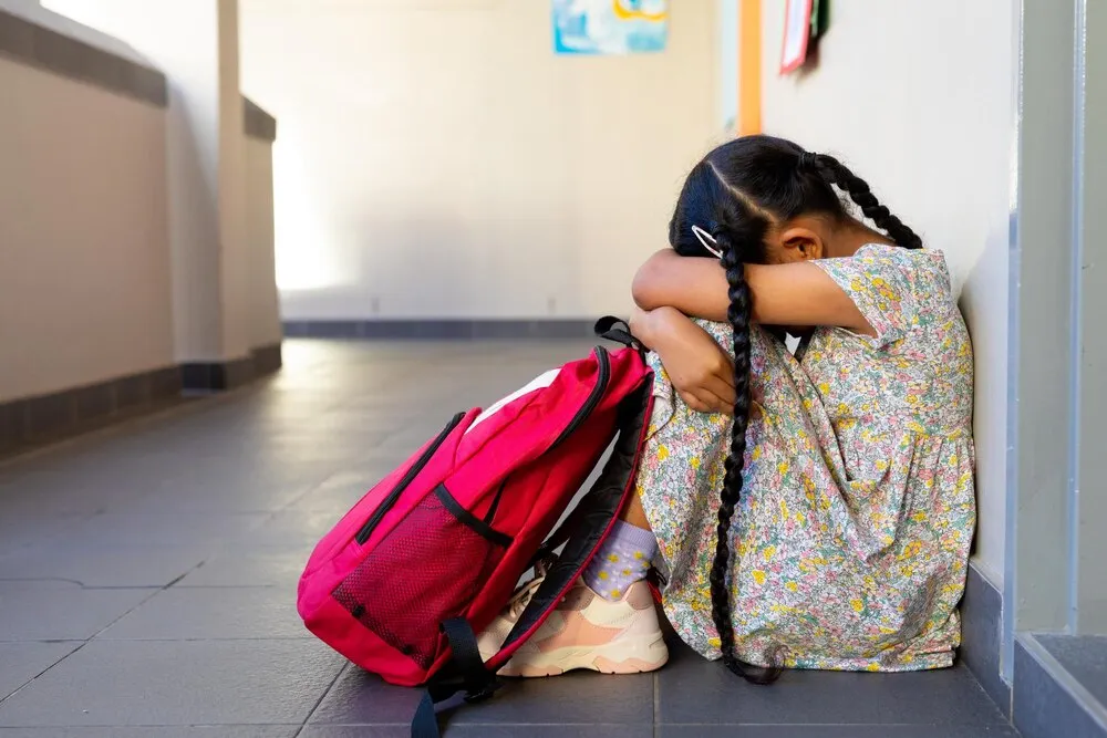 triste niña birracial con mochila sentada junto a la pared en el pasillo de la escuela primaria, espacio para texto