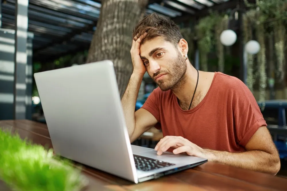 hombre joven y guapo cansado sentado con una laptop en una cafetería al aire libre, trabajando de forma remota o estudiando usando el wifi del parque