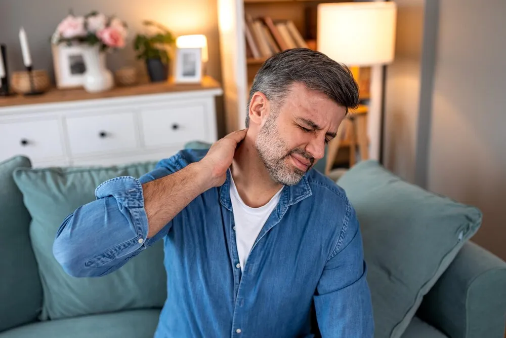 dolor de cuello y distensión muscular hombre adulto sentado en el sofá y sosteniendo el cuello dolorido con la mano concepto de salud y medicina hombre atractivo sintiéndose agotado y sufriendo de dolor cervical