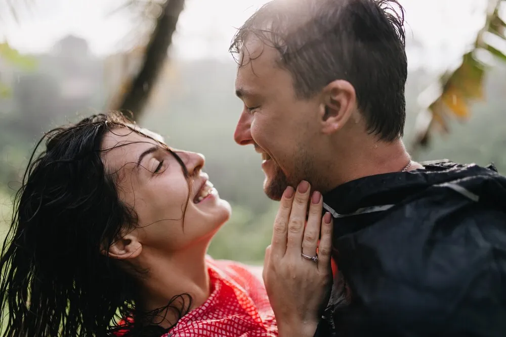 Hombre sonriente con impermeable mirando con amor a una mujer morena. Pareja romántica riendo, de pie en la naturaleza.