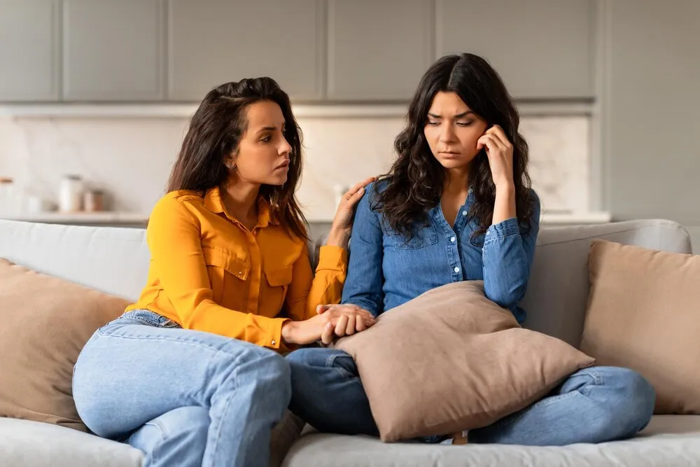 Mujer joven atenta consolando a una amiga deprimida sentadas en un sofá en interiores