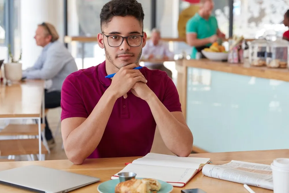 Toma de un joven escritor guapo y serio de etnia mixta que sostiene una pluma, hace una lista de pendientes en una libreta, toma café, pasa su hora de comida solo en una cafetería, ocupado con el trabajo.