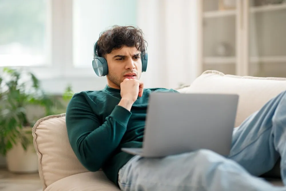 hombre árabe molesto con audífonos usando una laptop con problemas en interiores