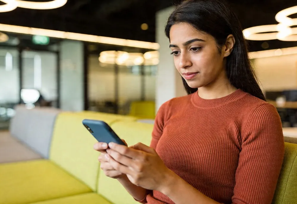 mujer india concentrada usando un smartphone en una oficina moderna en un sofá amarillo